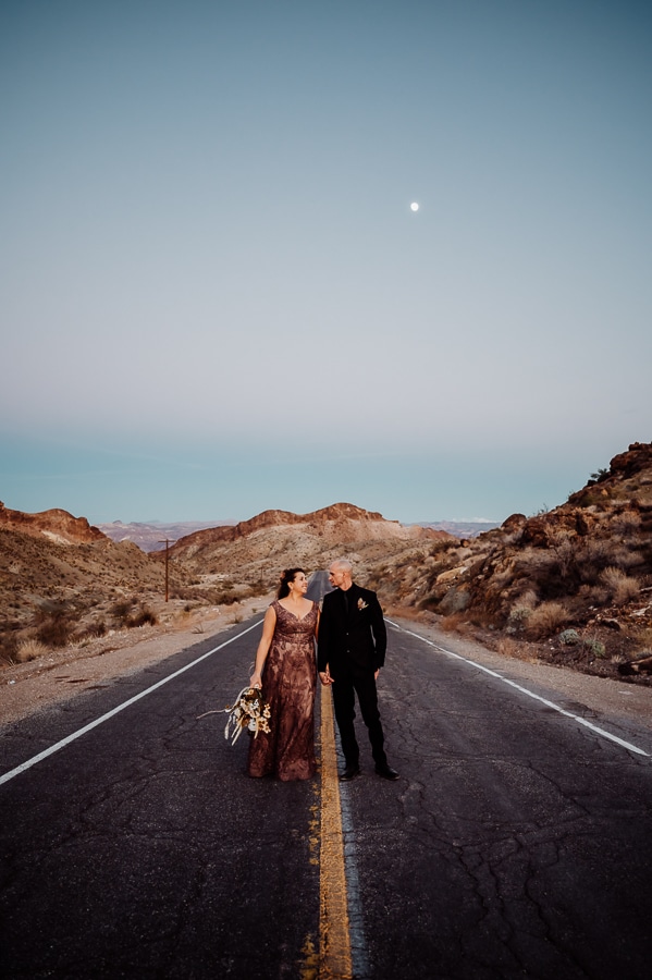 Bride and groom walking down the road at blue hour in front of moon in mountains