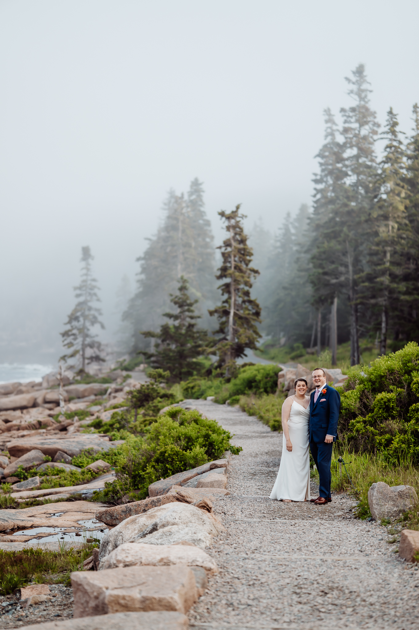 Bride and groom pose together on a gravel path lined with greenery and evergreens, with coastal cliffs and ocean mist in the distance at Acadia National Park.