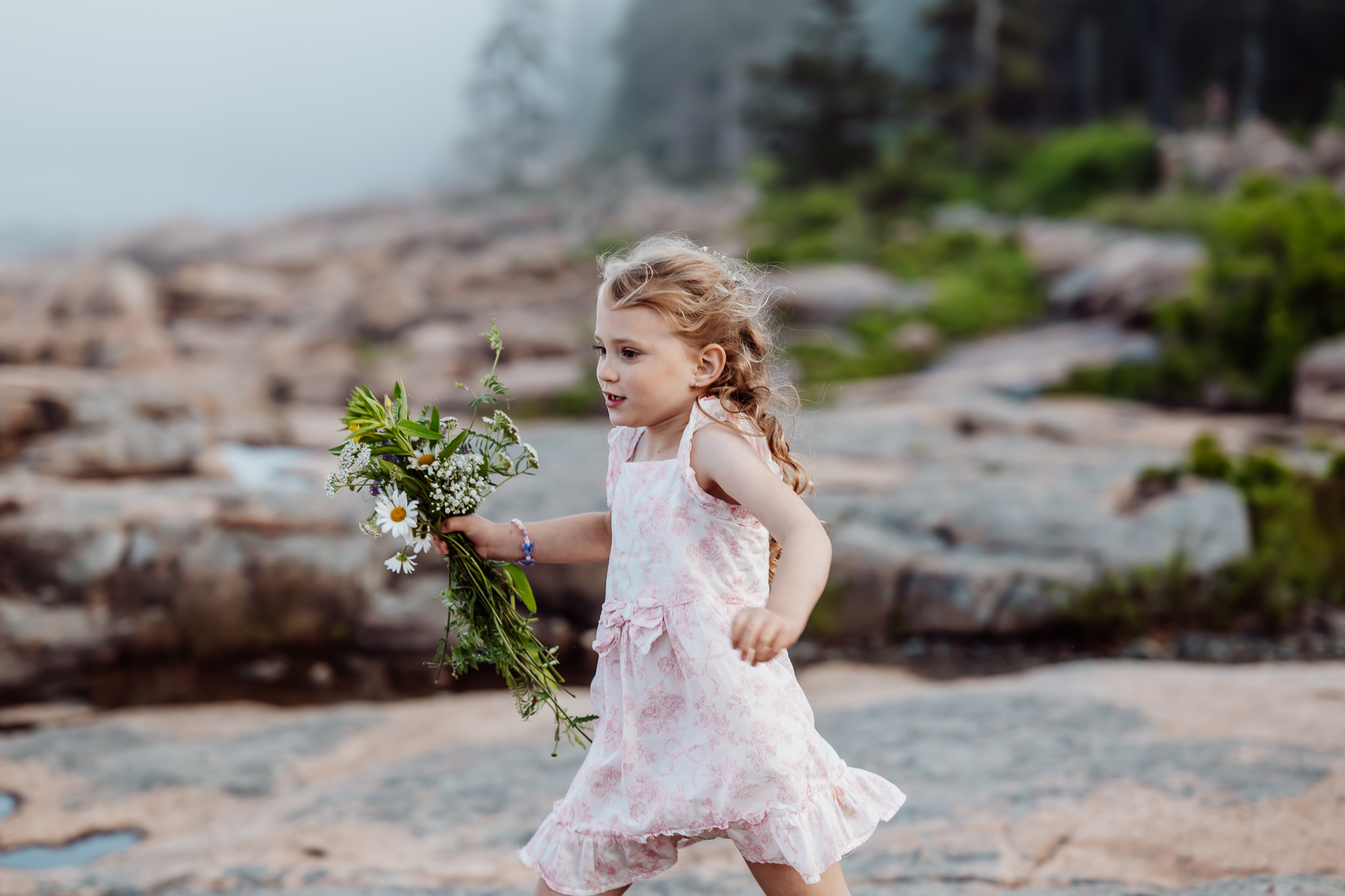 Young flower girl in a pink dress joyfully runs across the rocky shoreline, holding a wildflower bouquet with daisies and greenery, with forest and fog in the background.