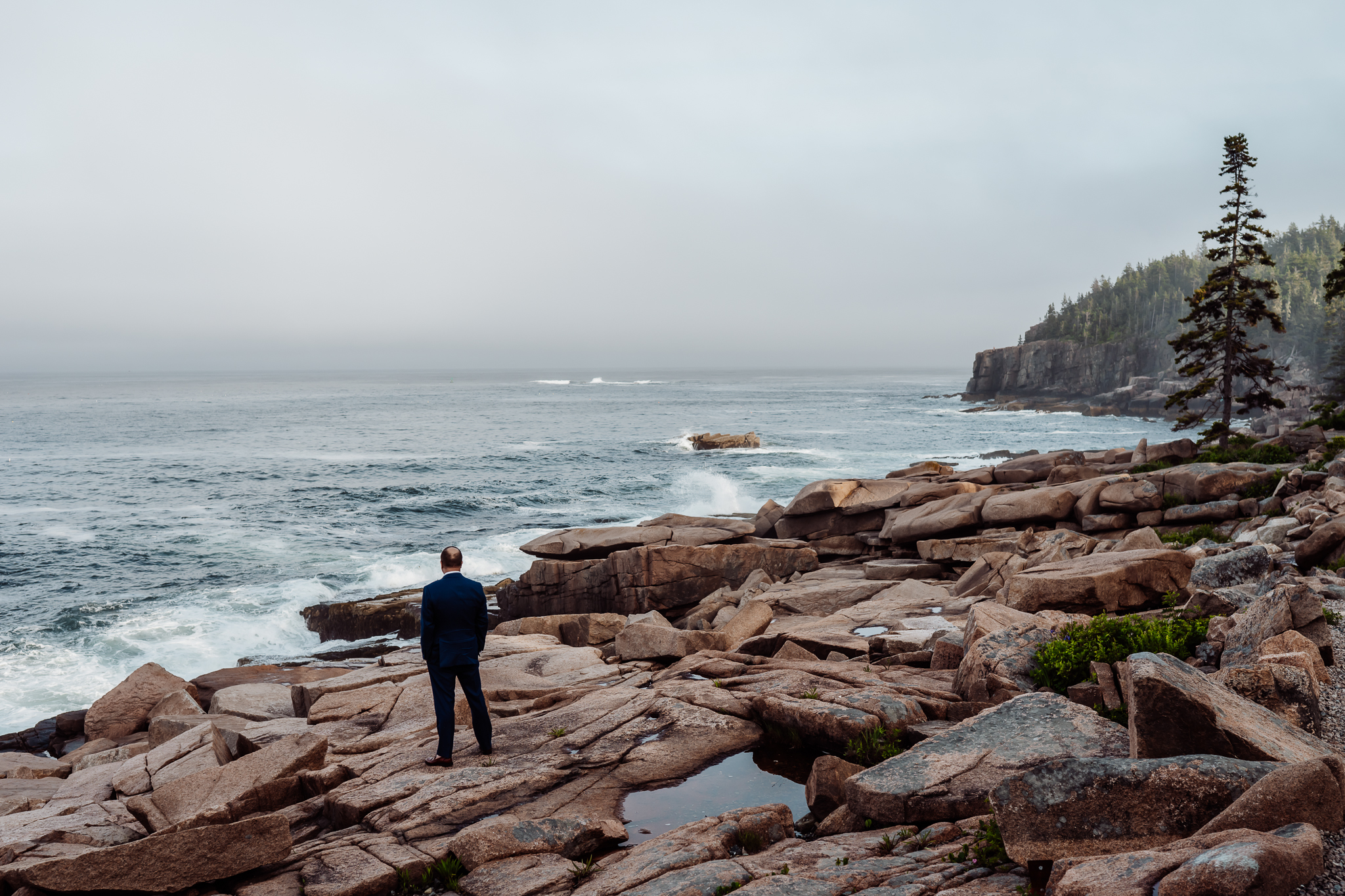 Groom stands alone on the rocky cliffs of Acadia National Park, gazing out at the vast Atlantic Ocean under a misty sky.