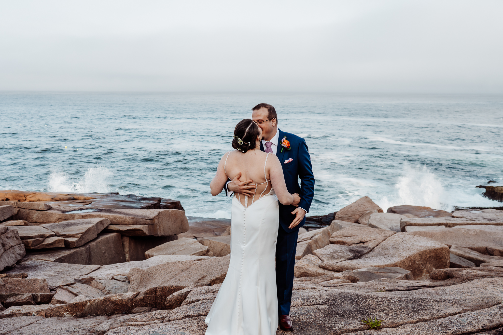 Bride and groom share a kiss on the rugged coastline, with waves crashing behind them and fog hovering over the ocean horizon.