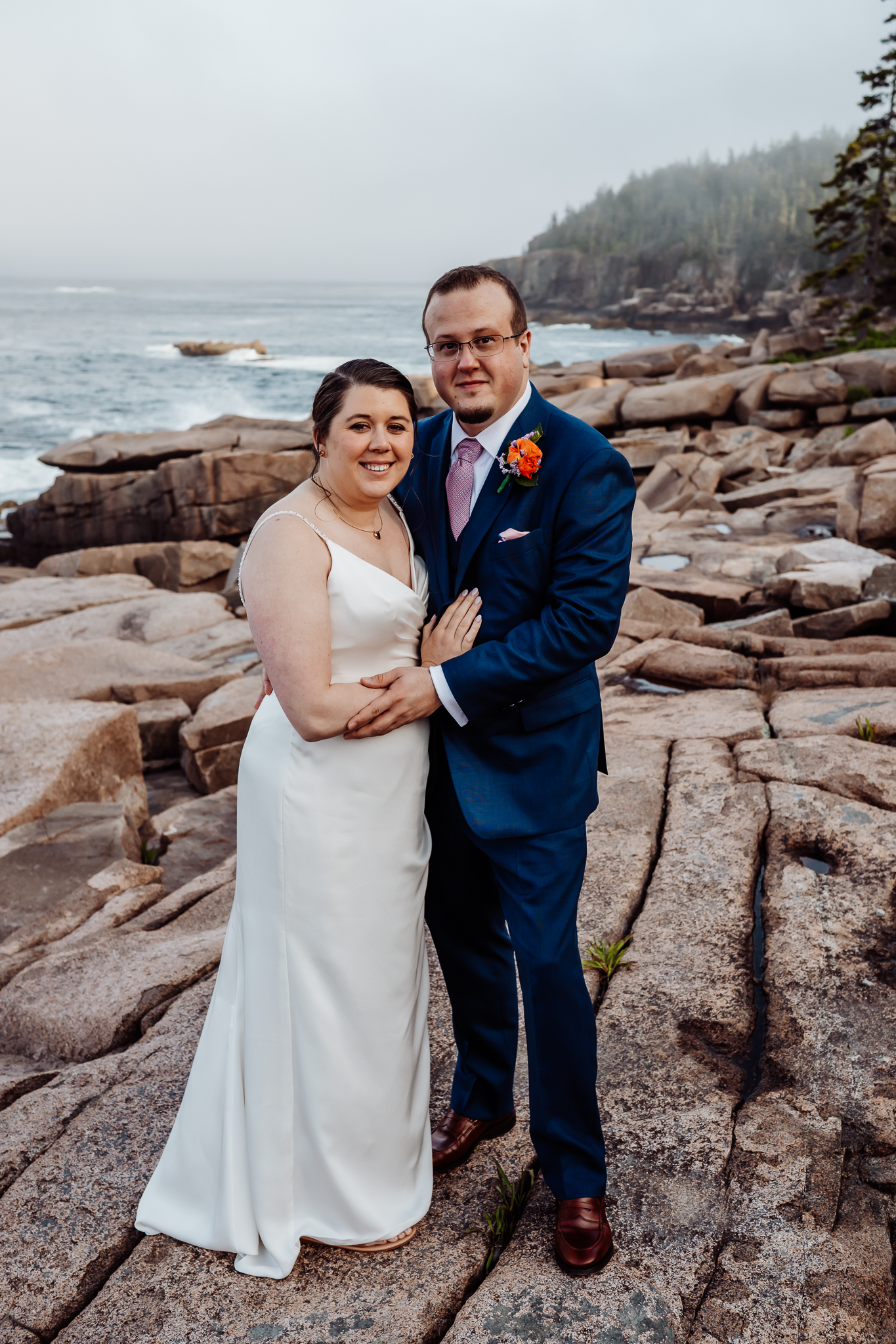 Bride and groom pose together on the rocky shore, smiling at the camera with the ocean and cliffside forest of Acadia behind them.