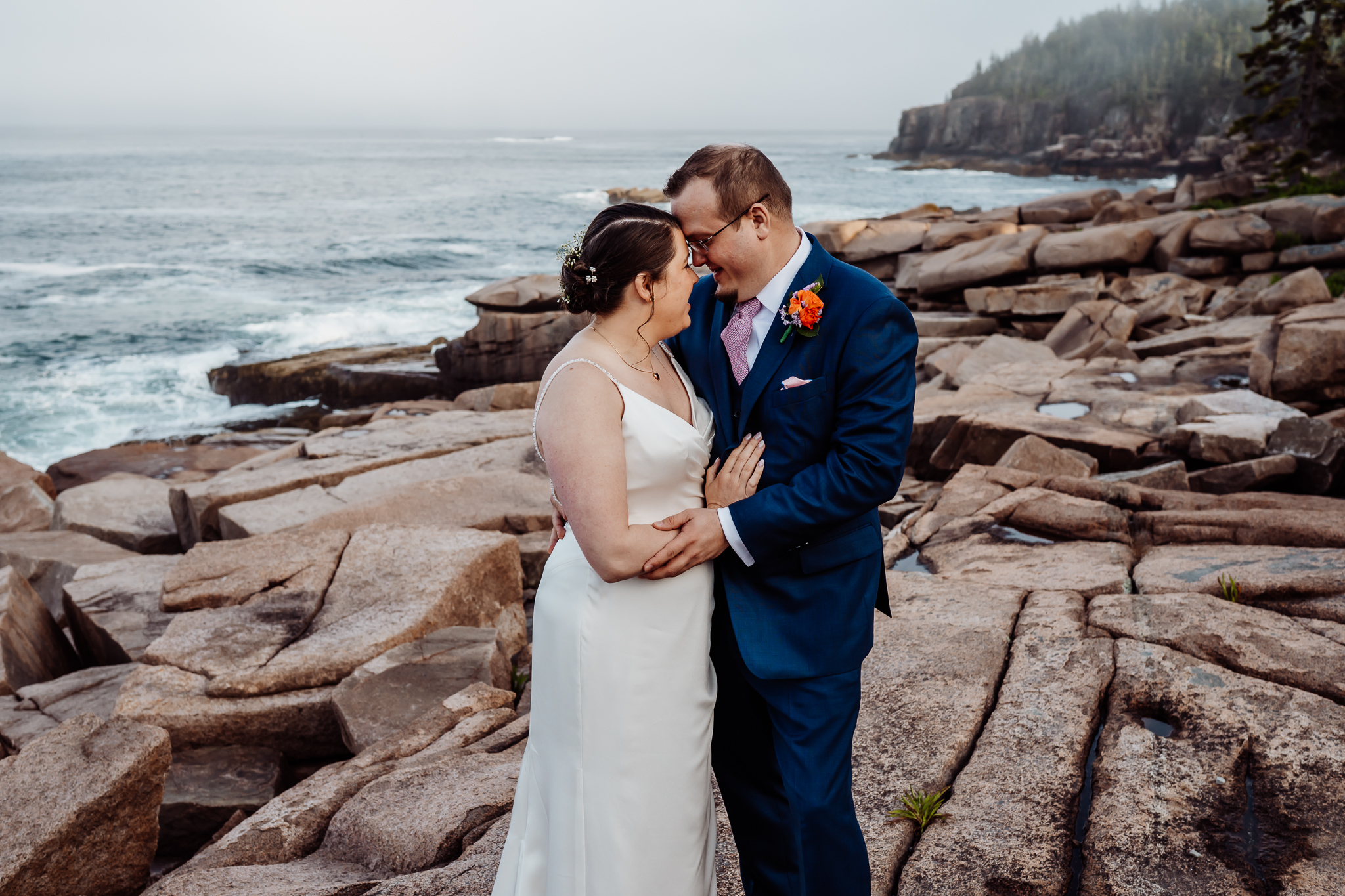 Couple shares an intimate moment on the rocky Maine coast, pressing their foreheads together with joyful smiles and ocean waves in the background.