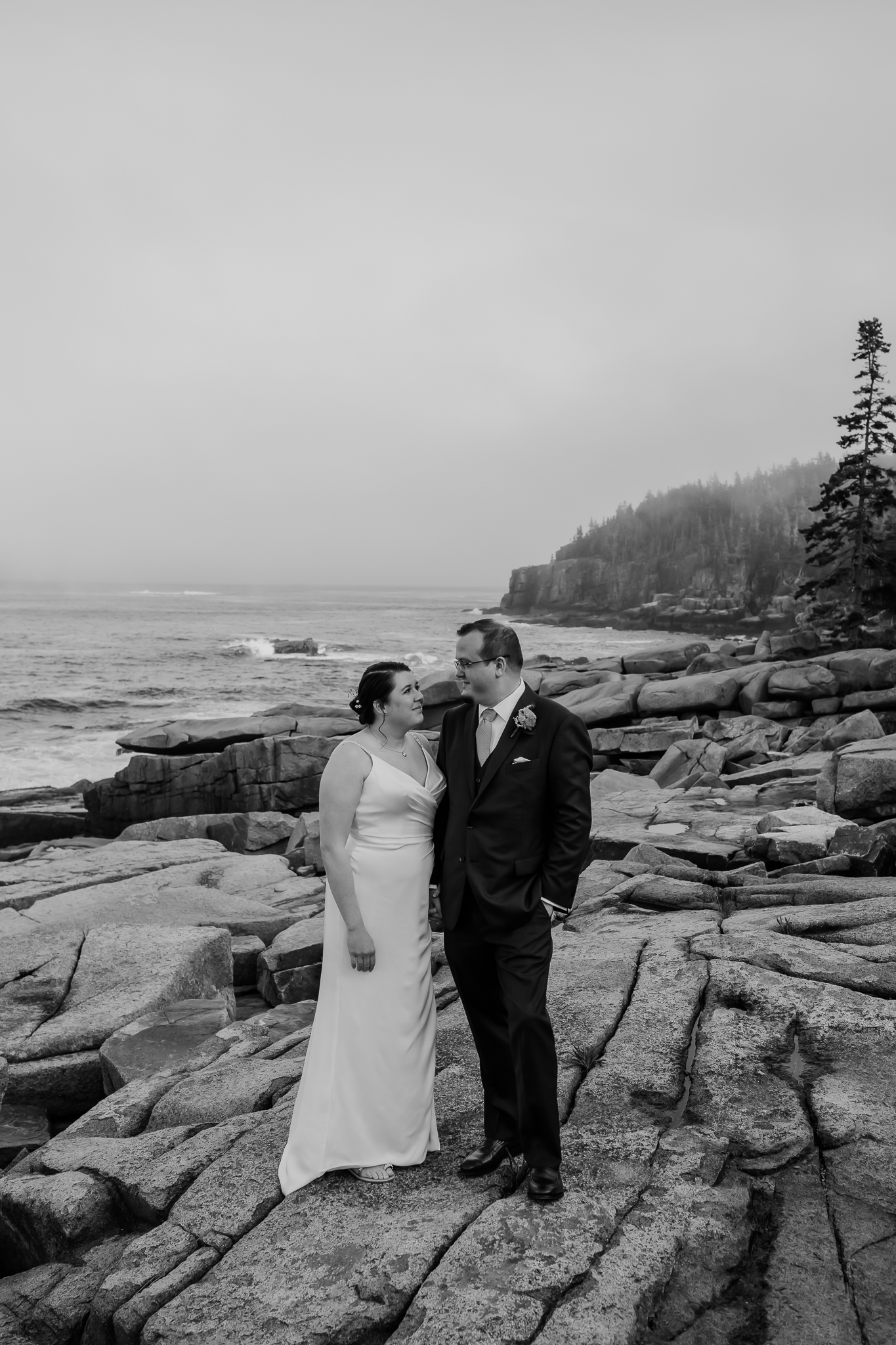 Black and white photo of a newlywed couple standing side by side on the rocky shoreline of Acadia, gazing at each other with the ocean stretching out behind them.