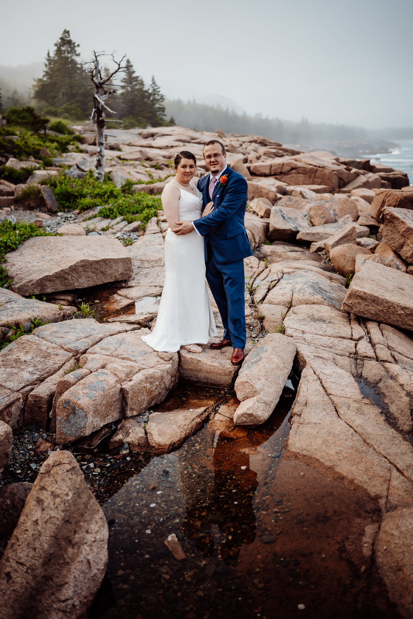 Bride and groom pose together among coastal rocks and greenery, looking directly at the camera with a misty forest behind them.
