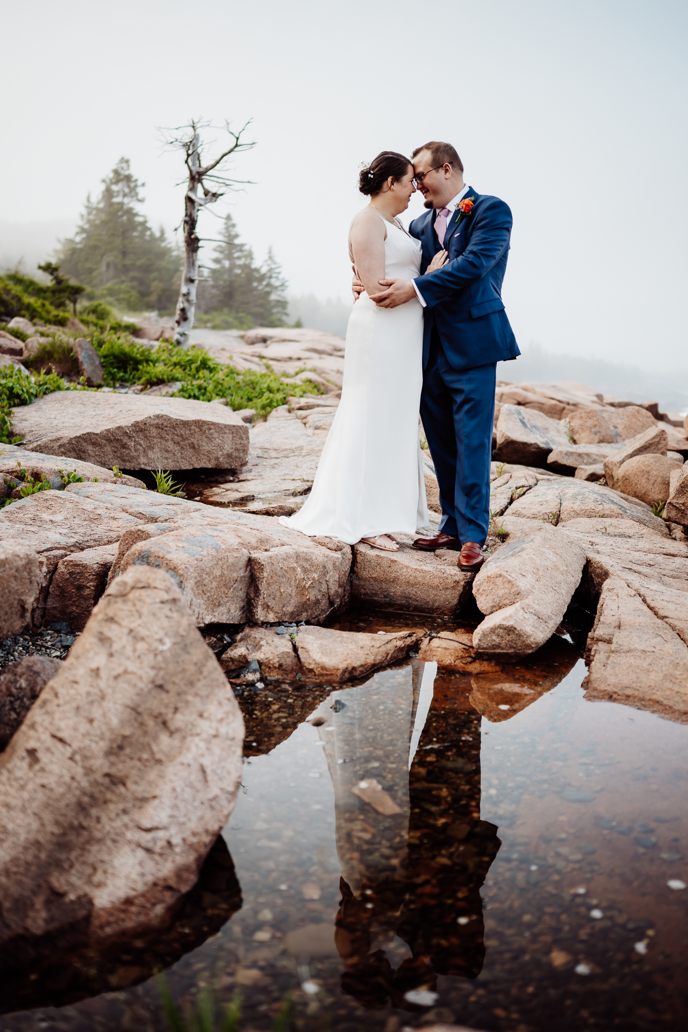 Newlyweds touch foreheads and smile, standing closely together above a tide pool that reflects their image, with foggy pine trees in the distance.