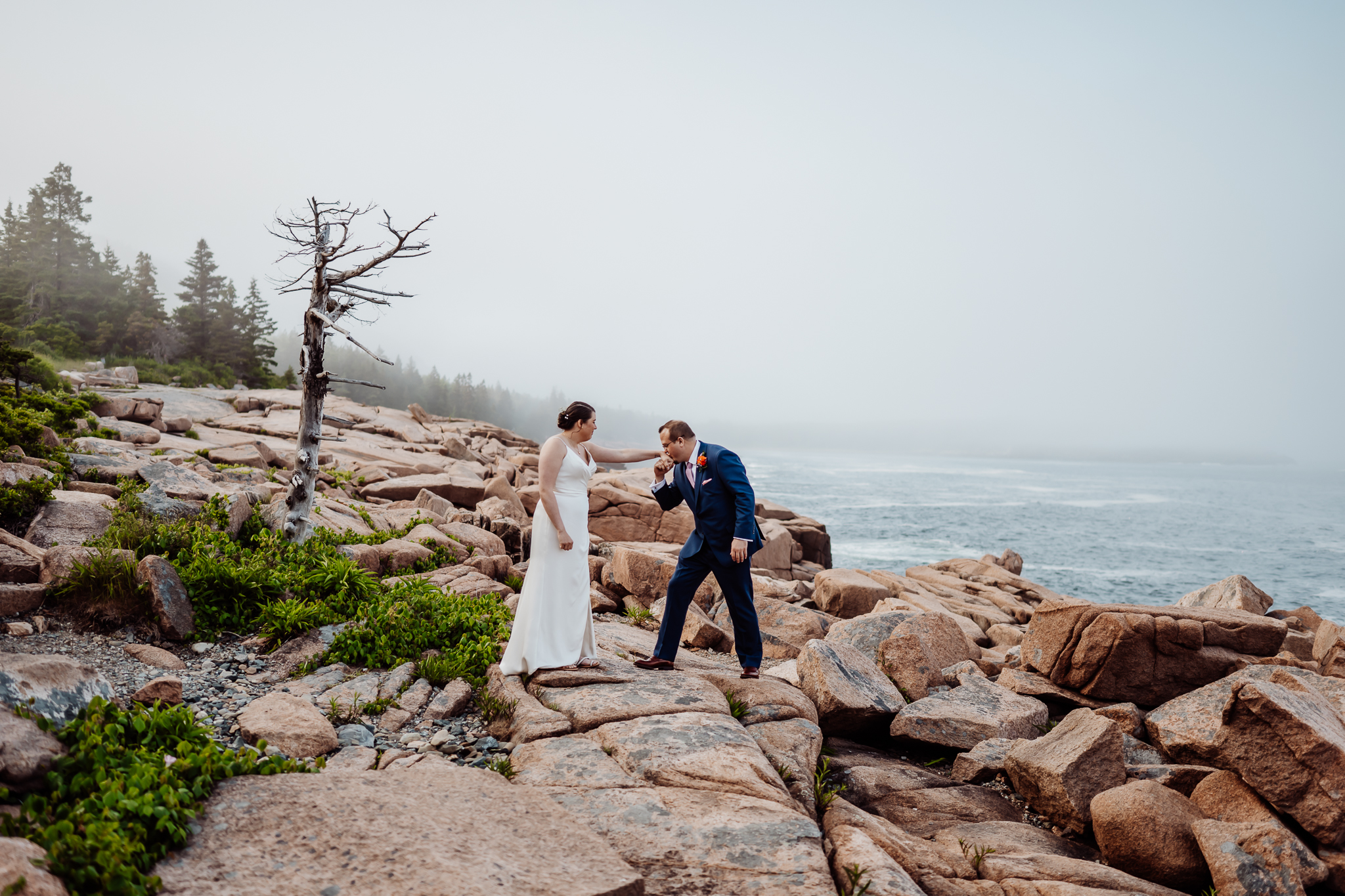 Groom kisses the bride’s hand as she smiles, standing on the rugged Maine coastline surrounded by ocean mist and wild greenery.