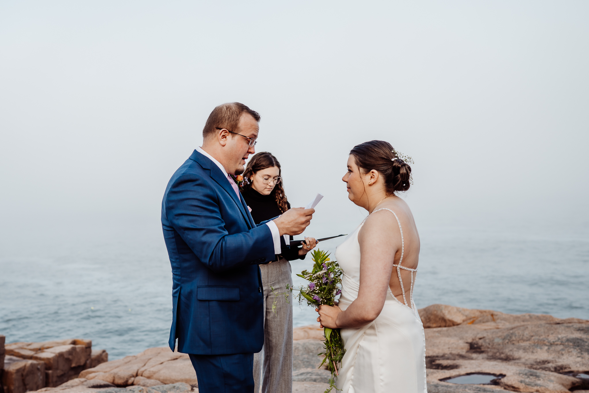Groom reads his vows to the bride during a coastal elopement ceremony in Acadia, with the officiant standing beside them on foggy oceanfront rocks.