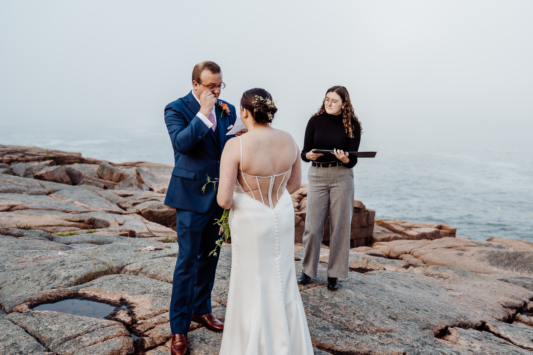 Groom wipes away tears while facing the bride during an emotional oceanside ceremony, with an officiant standing nearby on the rocky cliffs of Acadia National Park.