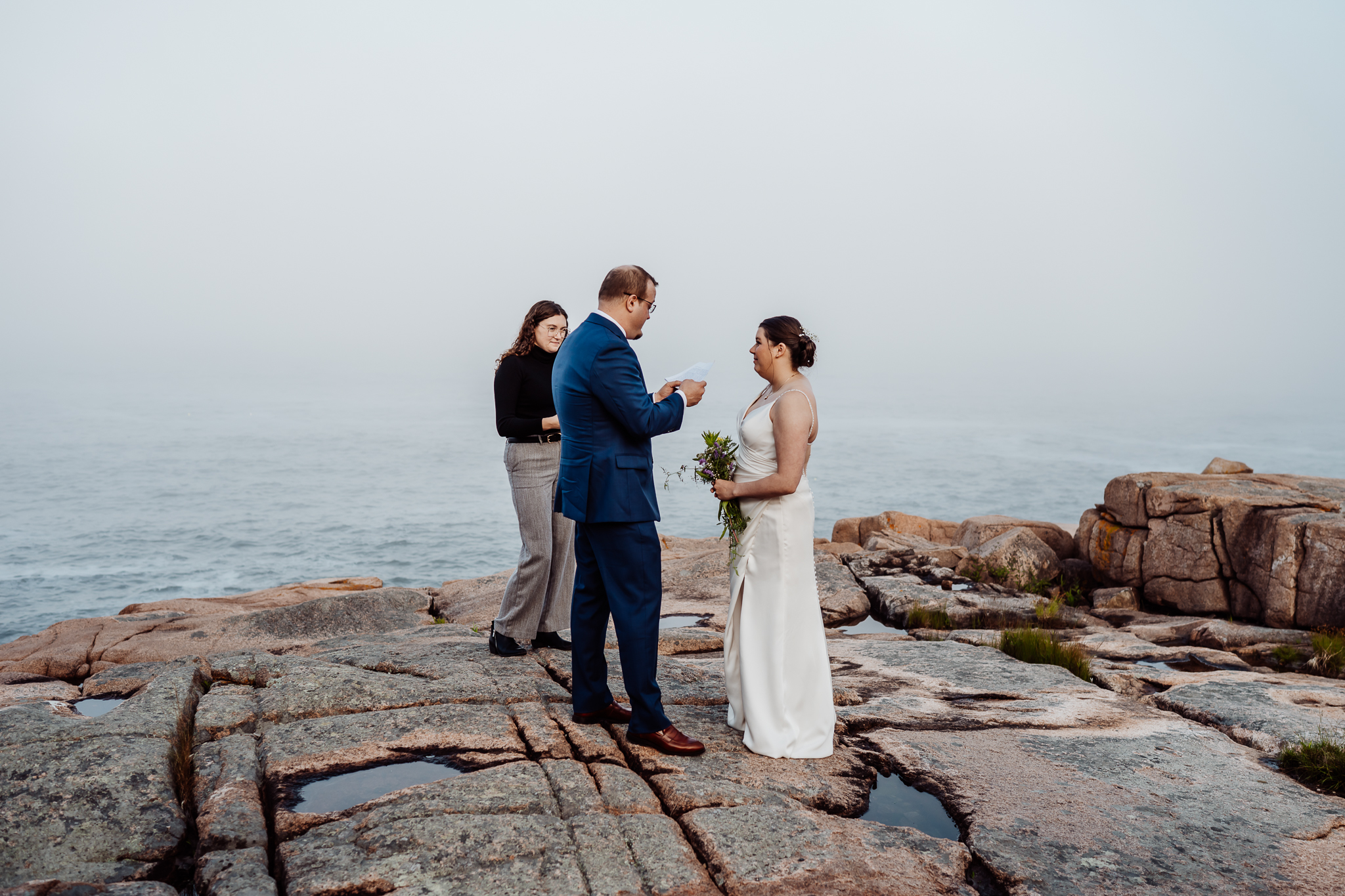 Bride and groom exchange heartfelt vows on the rugged shoreline of Acadia National Park, with the ocean and their officiant behind them in the morning fog.
