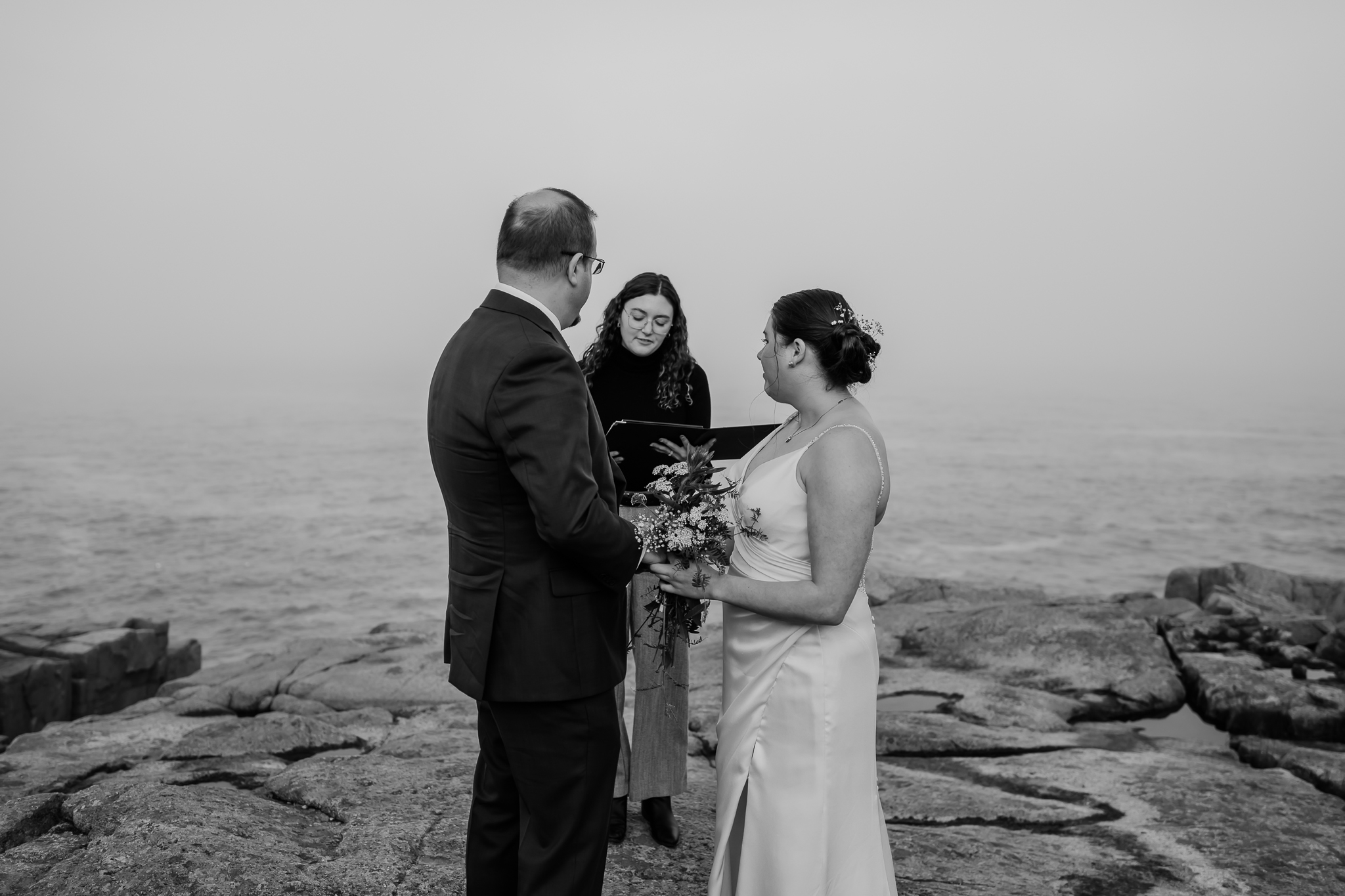 Black and white photo of an intimate wedding ceremony by the sea, as the officiant reads vows to the bride and groom on the rocky shoreline.