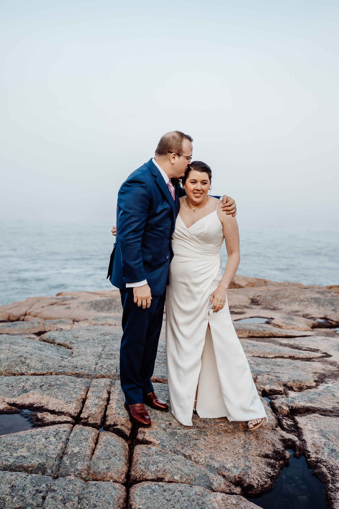 Groom kisses the bride on the forehead as they stand embraced on the rocky coastline, with misty skies and the Atlantic Ocean behind them.