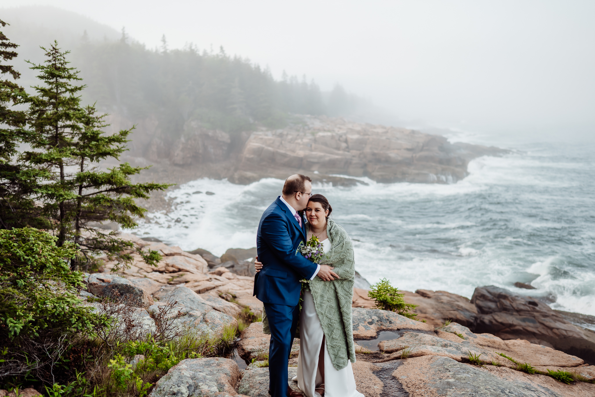 Bride wrapped in a green blanket and holding a bouquet, nestled into the groom’s chest as they embrace on a foggy cliff overlooking the crashing waves of Acadia National Park.