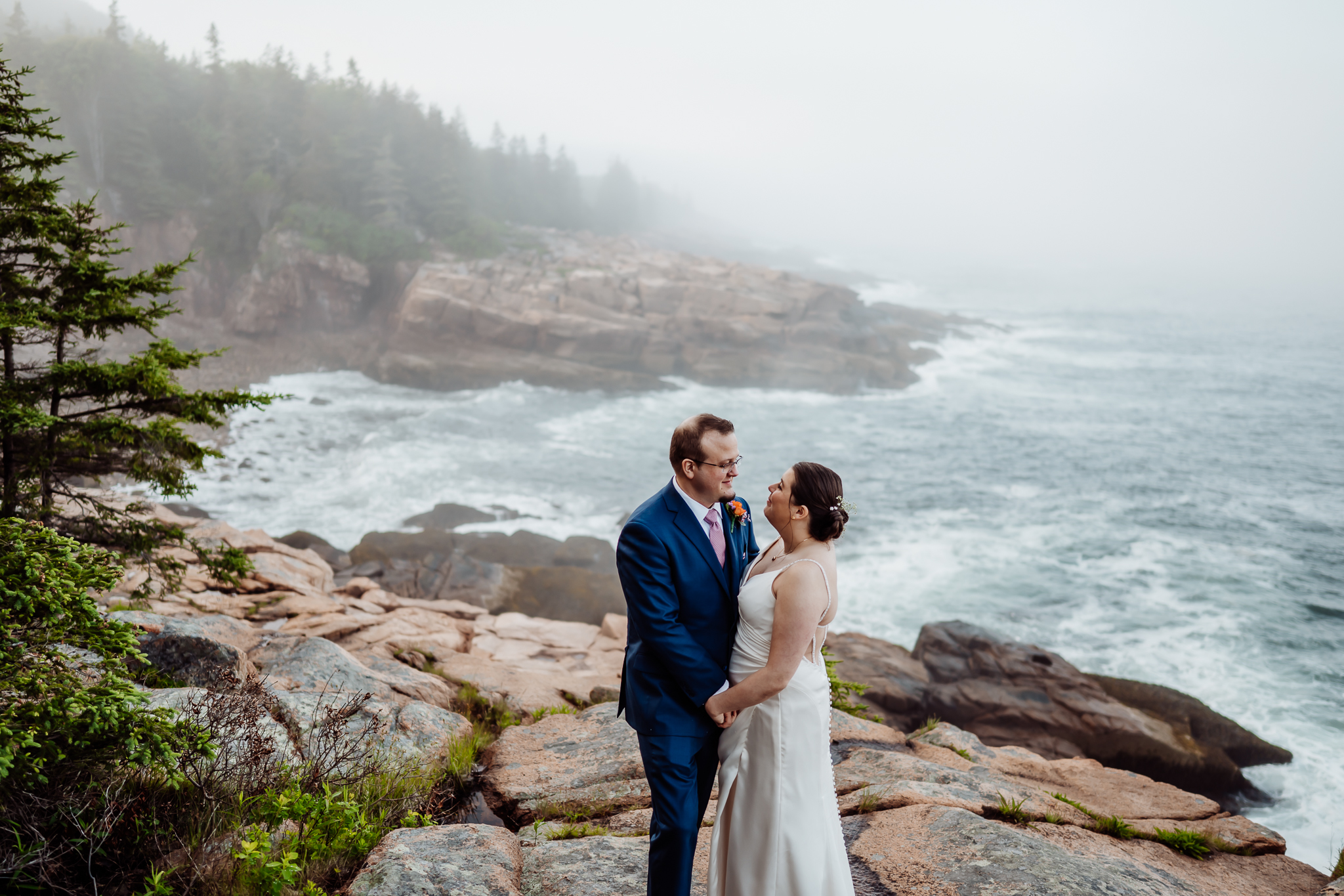 Newlyweds share a tender moment while holding hands on a foggy coastal cliff in Acadia National Park, with dramatic waves and evergreens in the background.