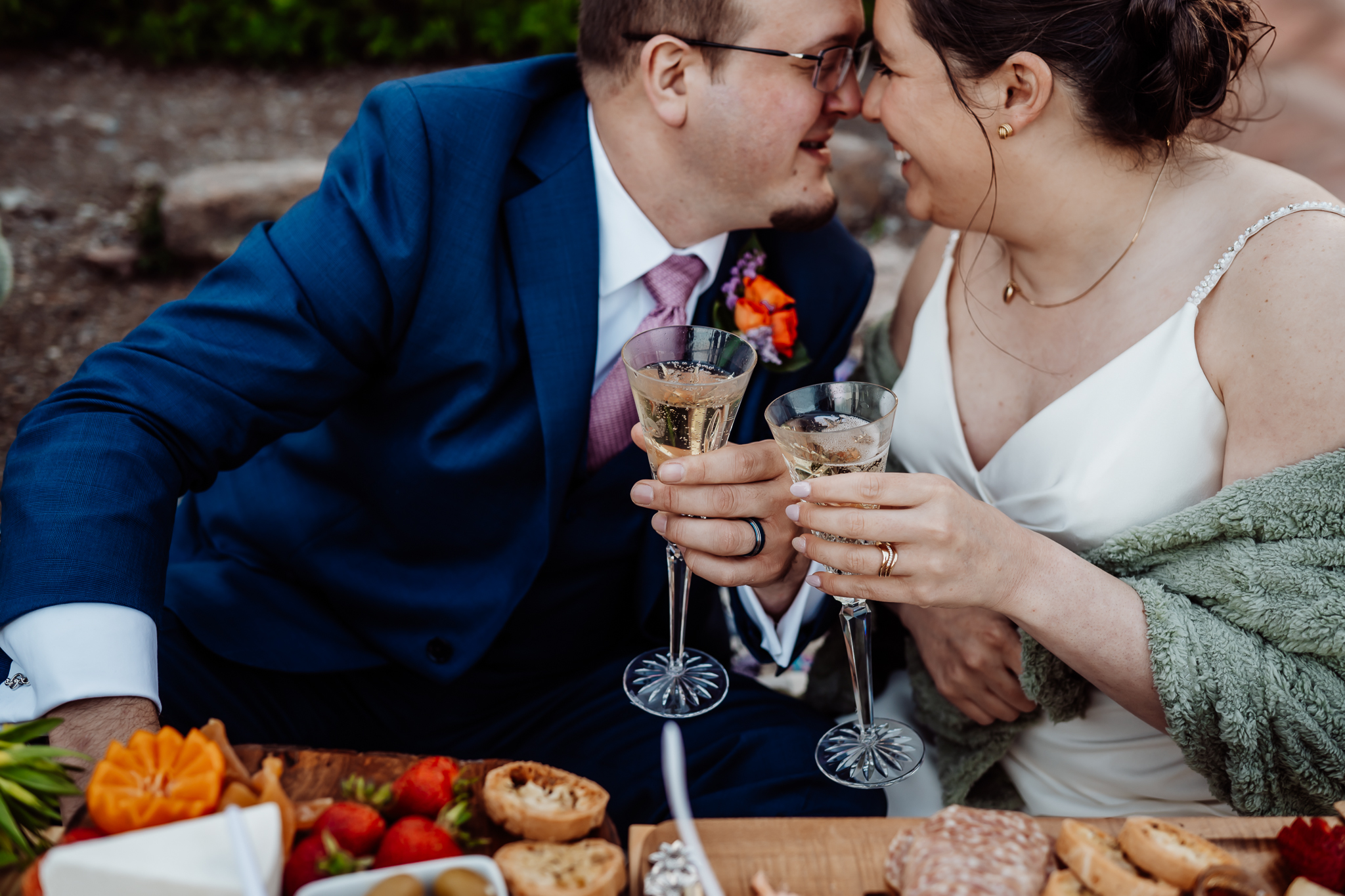Close-up of a newlywed couple toasting with champagne flutes during an intimate picnic, smiling and touching noses in joyful celebration.