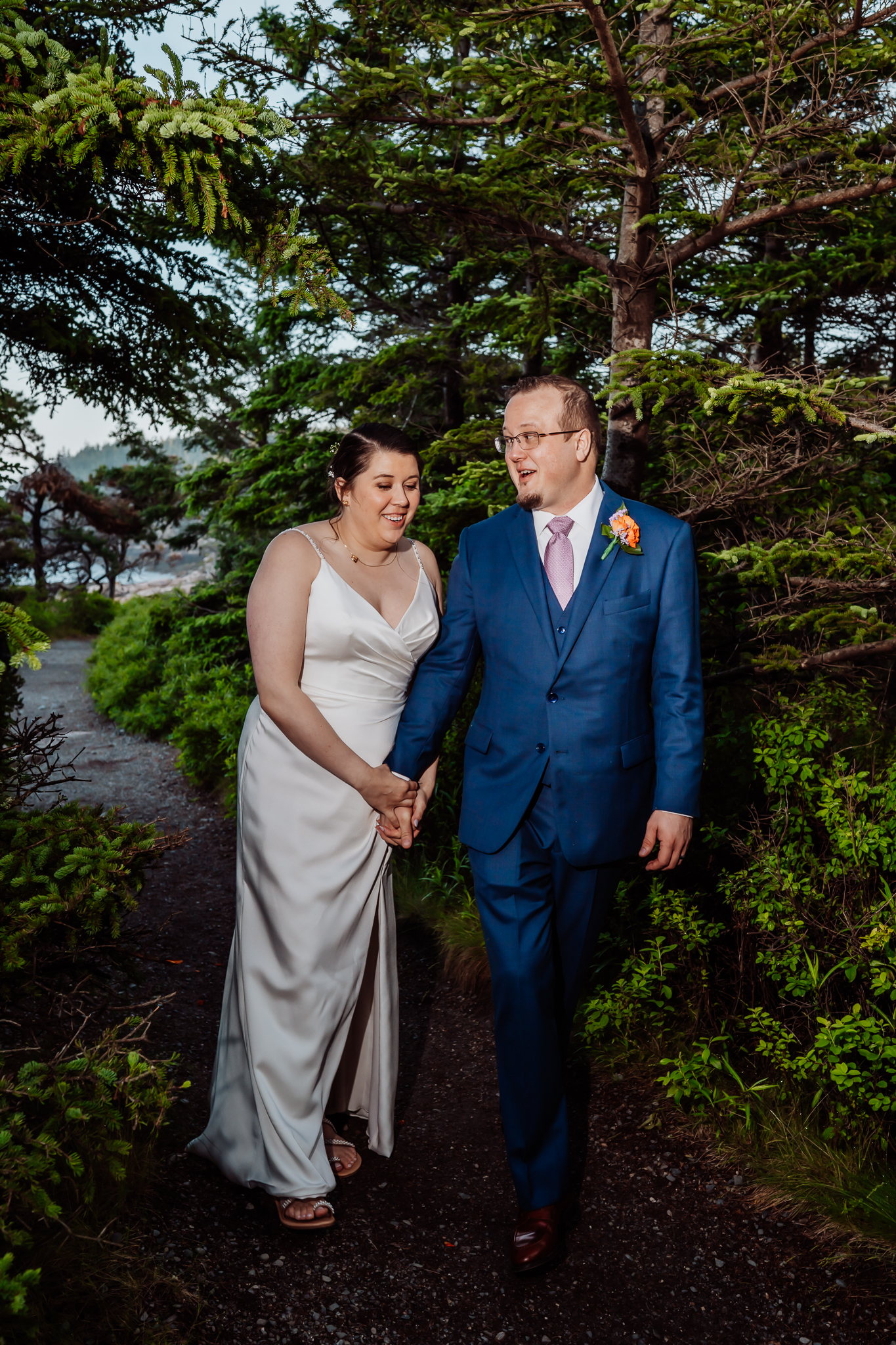 Bride and groom walking hand-in-hand down a forested path, laughing and smiling under lush evergreen trees in Acadia National Park.