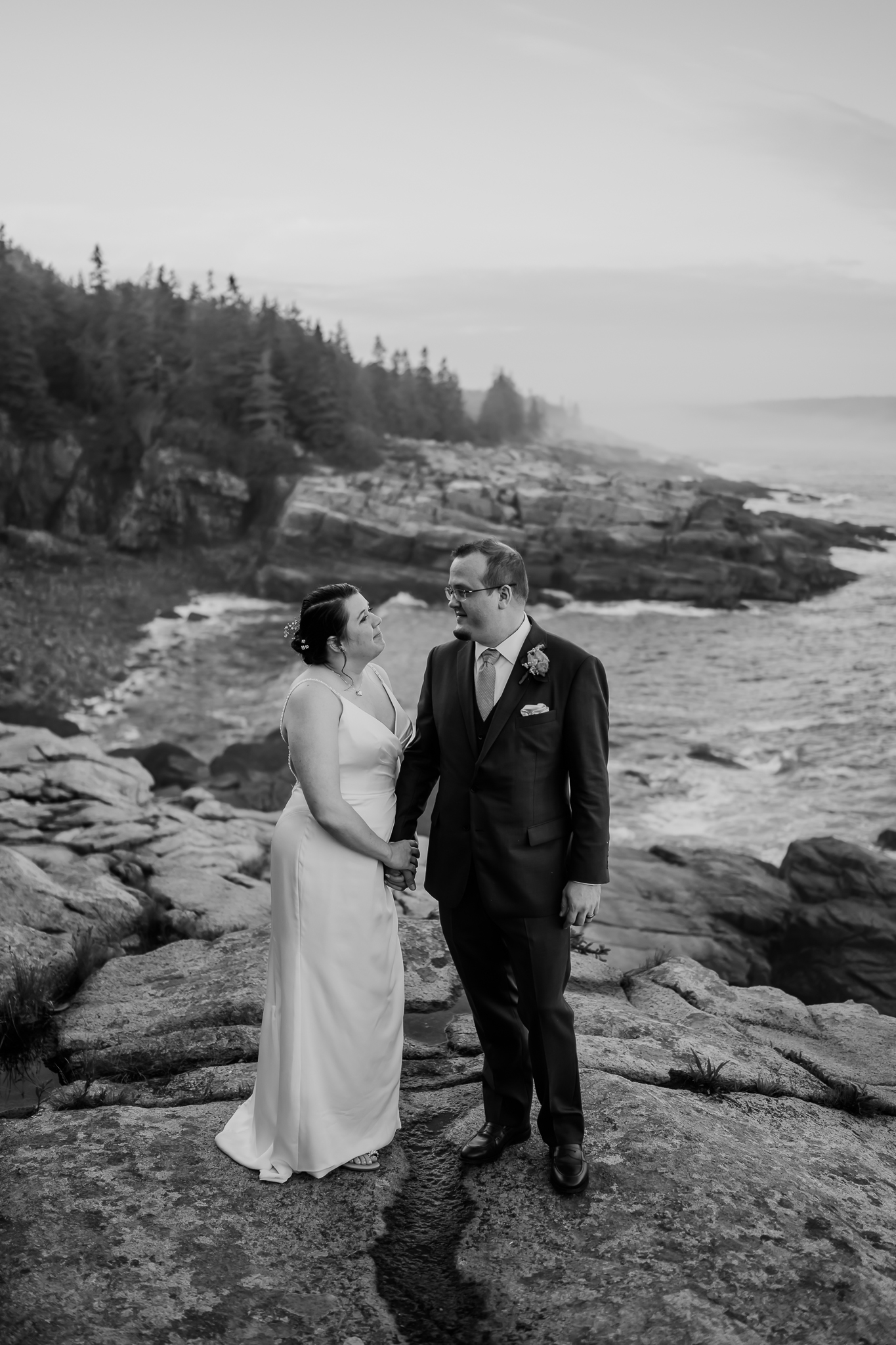 Black and white image of a newlywed couple holding hands and gazing at each other, standing on coastal rocks with the ocean and pine forest behind them.