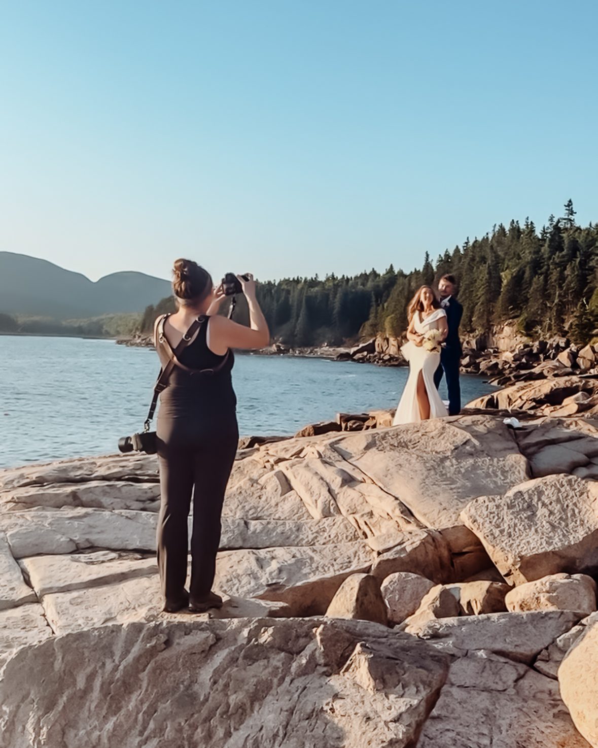 A wedding photographer stands on rocky cliffs in Acadia National Park, photographing a bride and groom embracing by the ocean with forested mountains in the background.