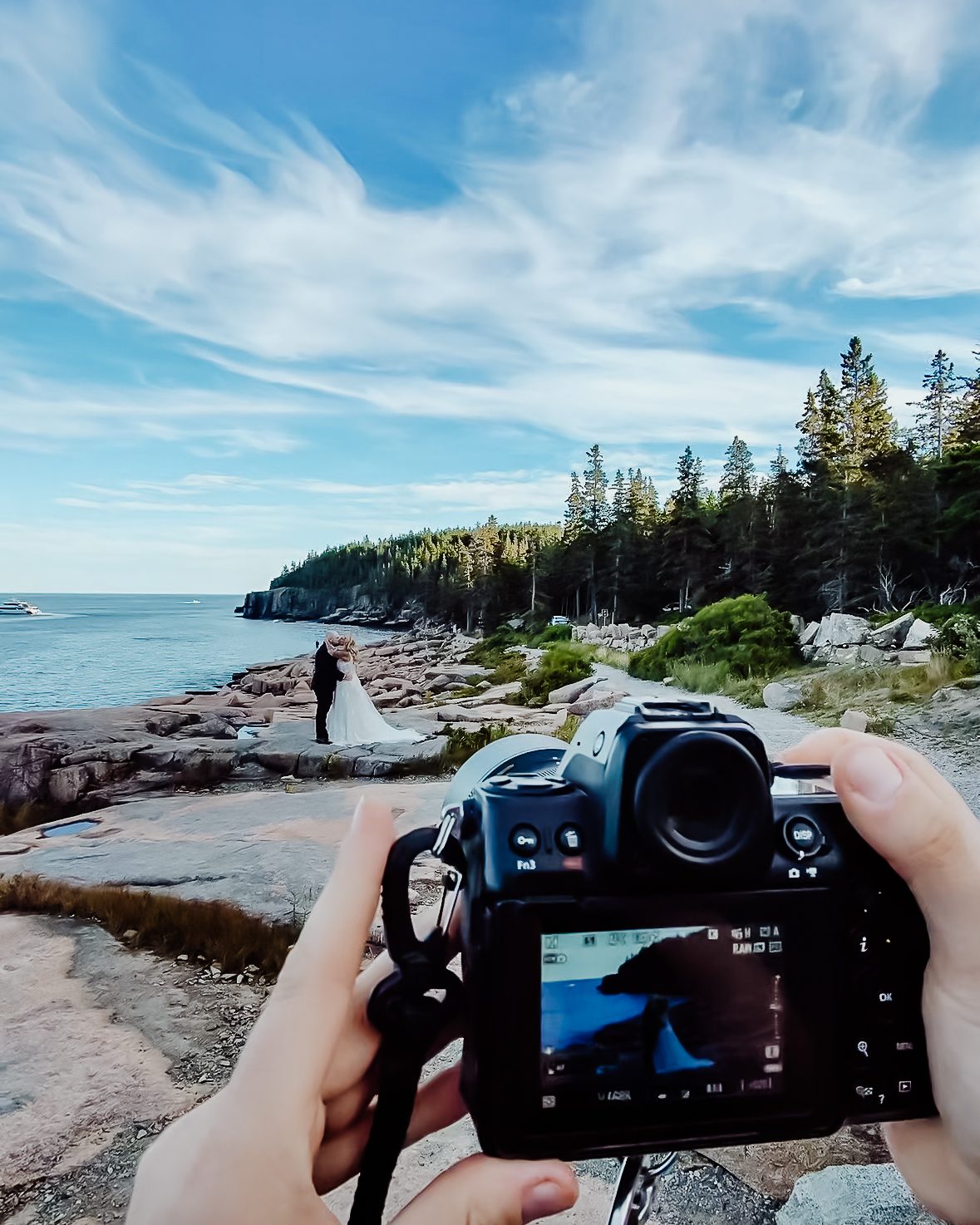 Close-up of a camera screen showing a bride and groom kissing on the rocky coastline of Acadia National Park, with the couple visible in the distance against the ocean and pine trees.