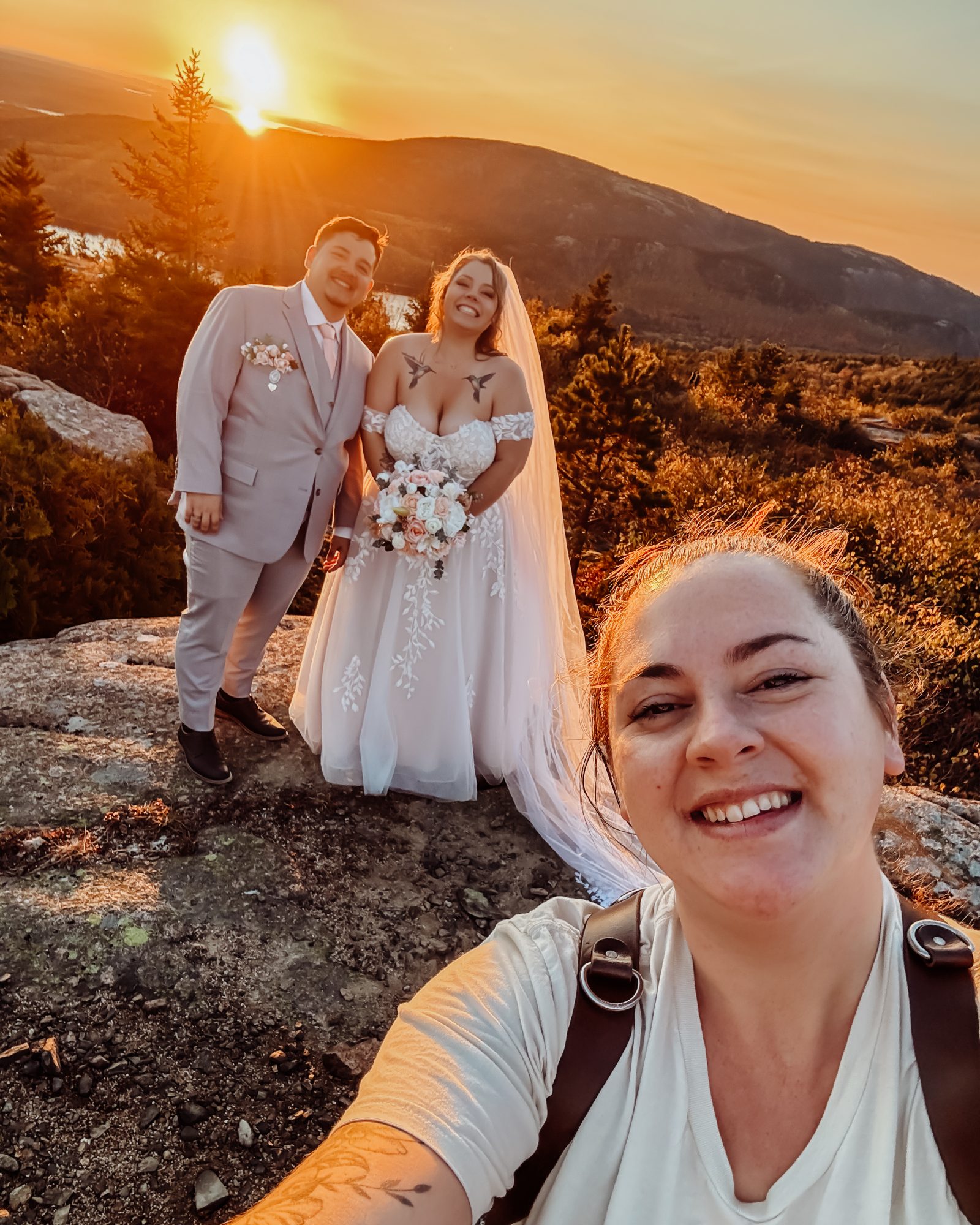 Behind the scenes of a smiling wedding photographer takes a selfie with a newlywed couple at sunset on top of a mountain in Acadia National Park, the sky glowing orange as the sun sets behind the peaks.
