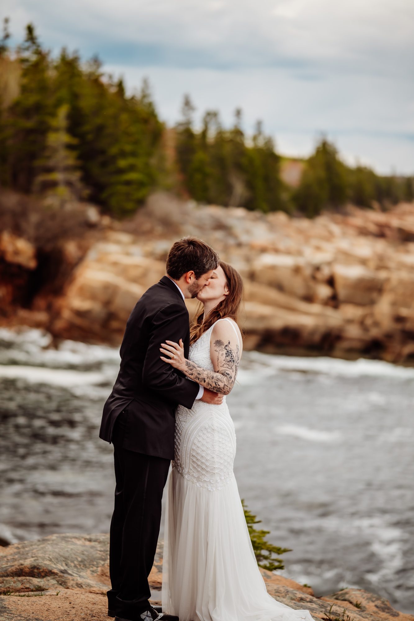 Couple kissing on the rocky coastline in Acadia National Park with ocean waves and evergreen cliffs in the background