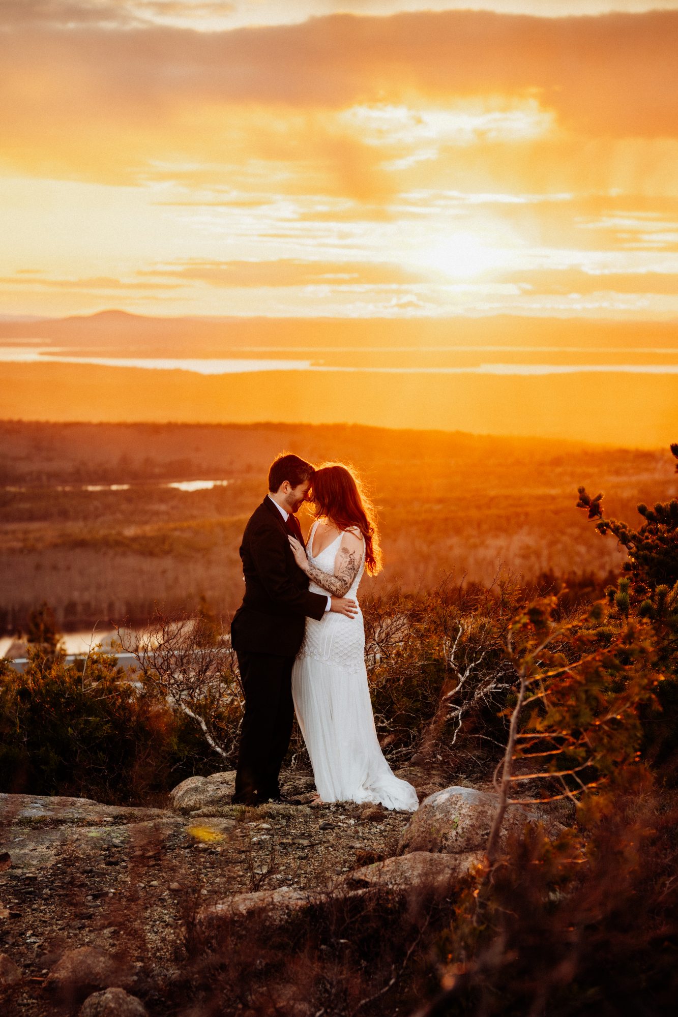 Newlywed couple sharing an intimate sunset moment on a mountaintop overlook in Acadia National Park with glowing golden light across the landscape