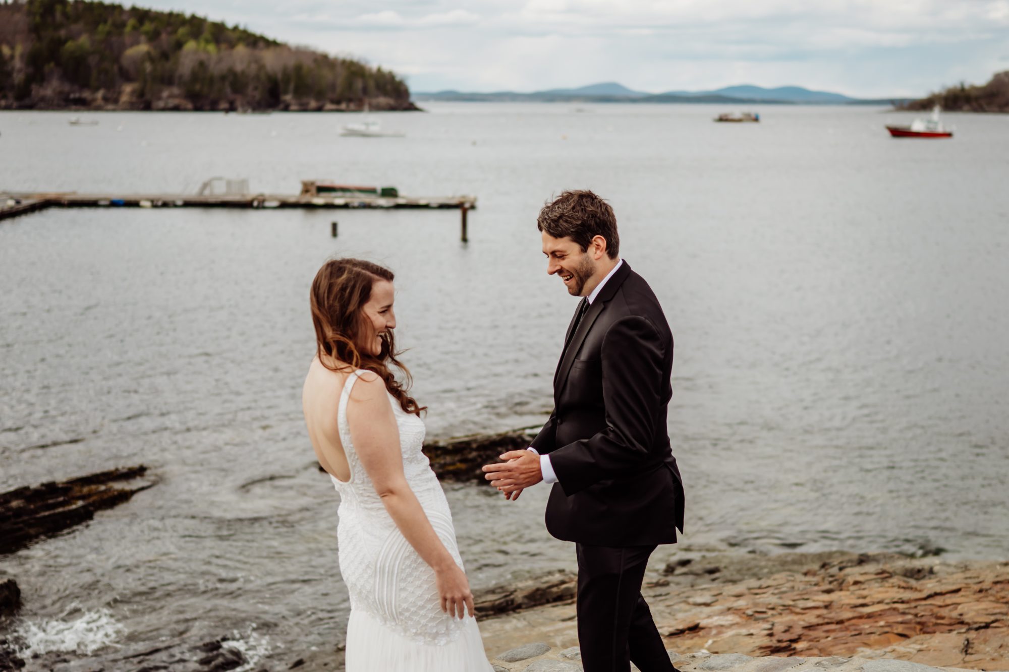 Joyful newlywed couple laughing together along the rocky shoreline near Frenchman Bay in Bar Harbor, Maine
