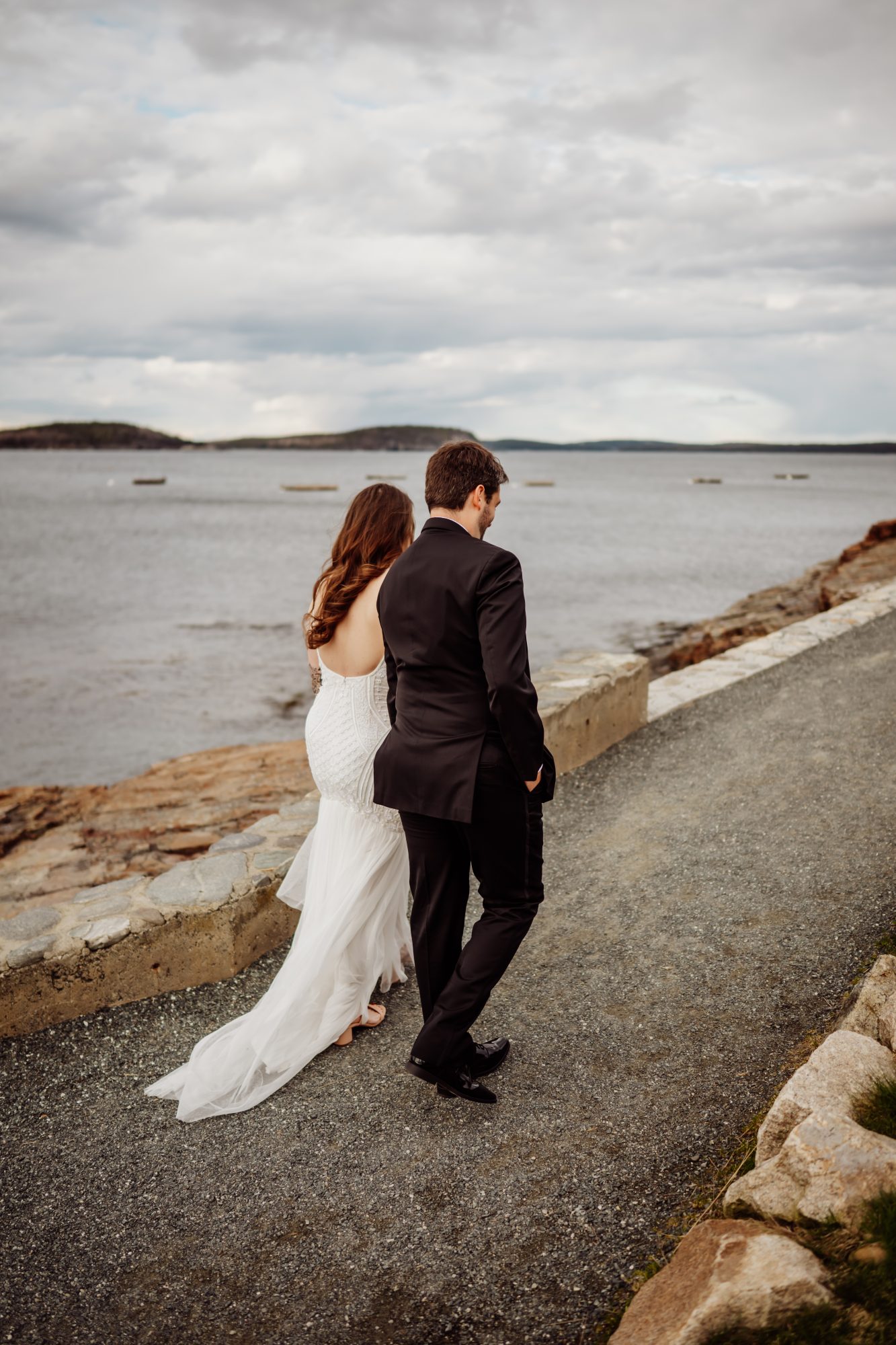 Windswept bride and groom walking together along the scenic coastal road near Frenchman Bay in Bar Harbor Maine