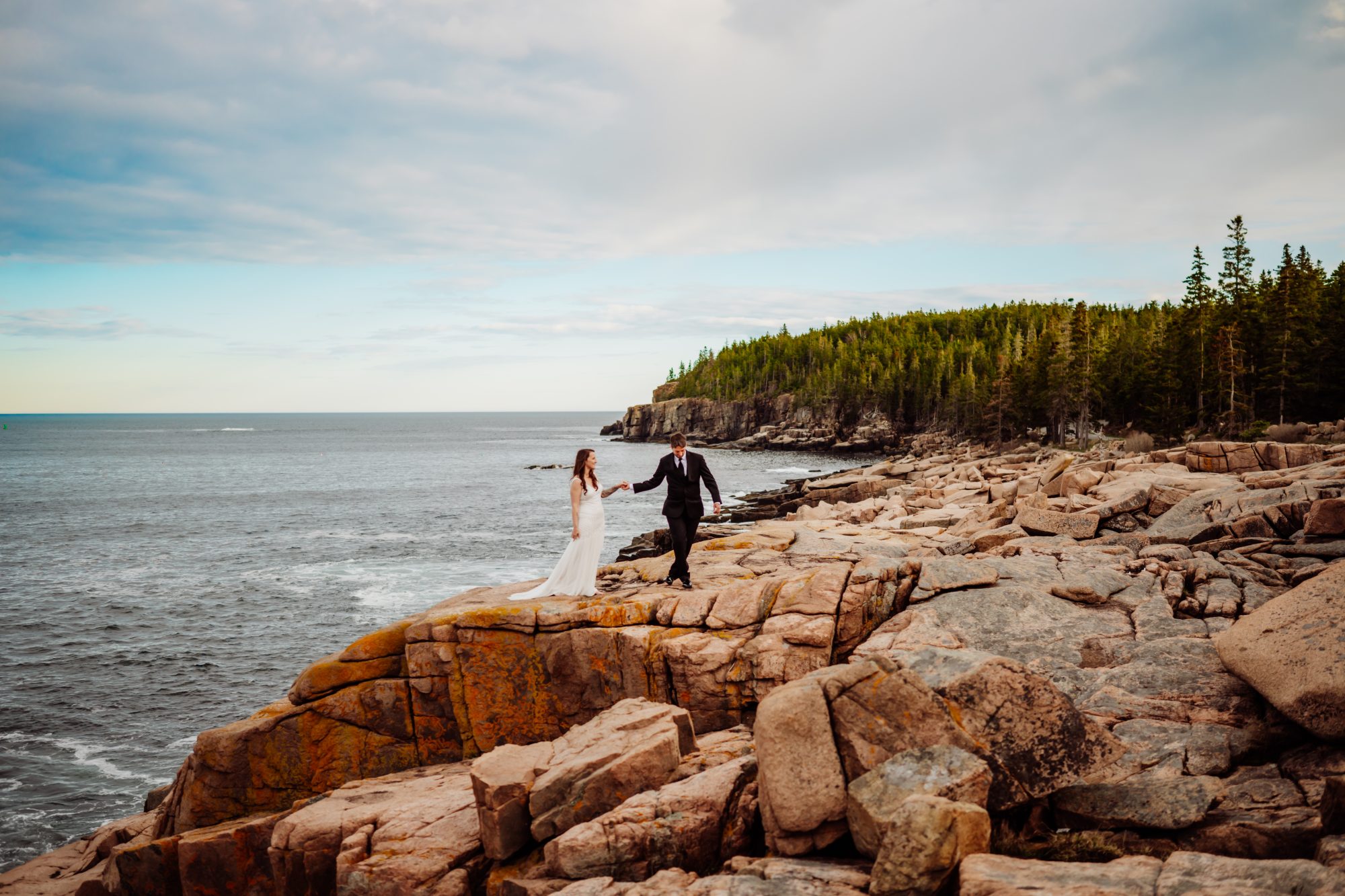 Bride and groom walking hand in hand along the granite cliffs of Acadia National Park with scenic coastal views