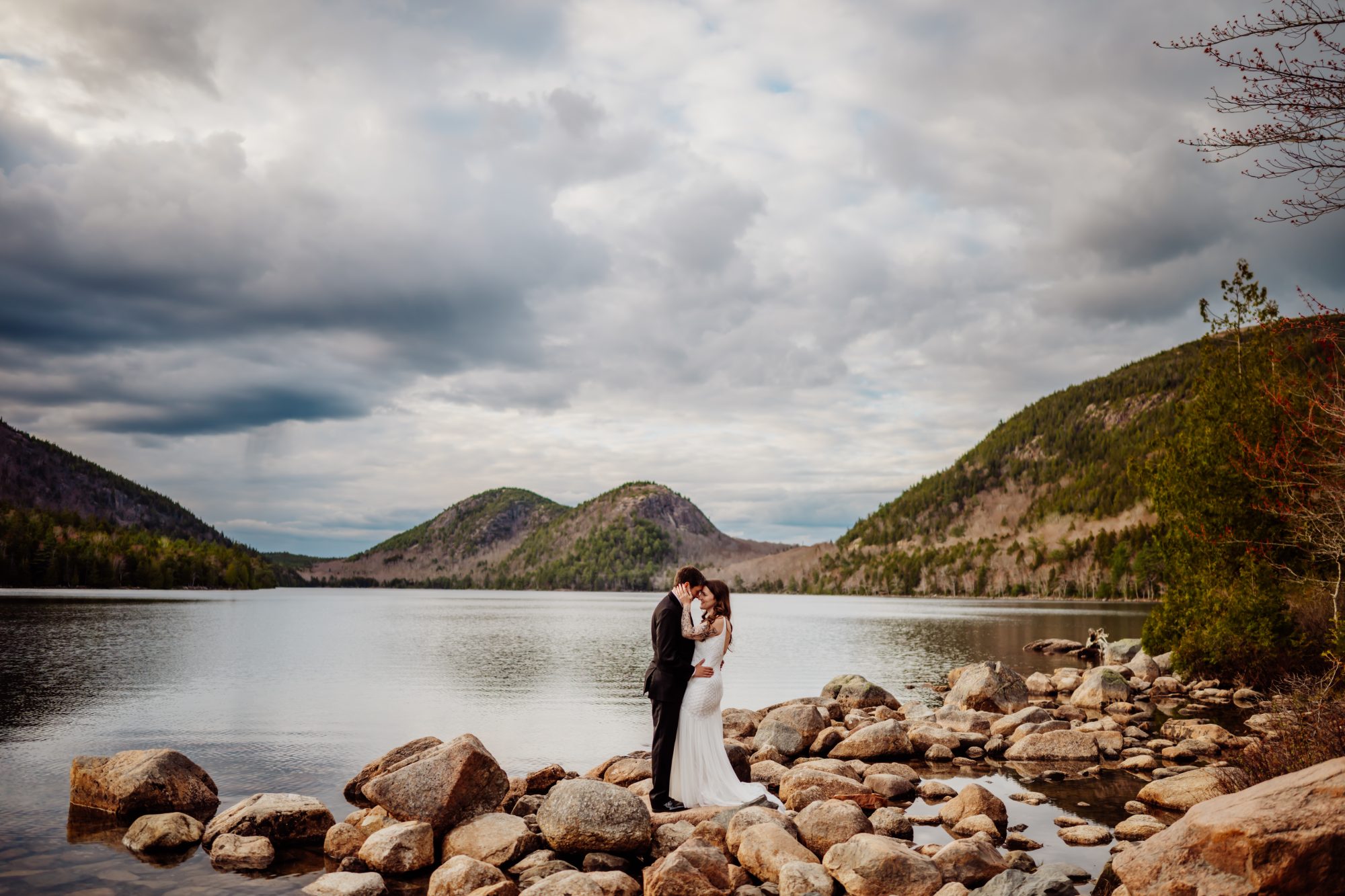 Wide scenic photograph of an eloping couple embracing beside Jordan Pond under dramatic clouds in Acadia National Park