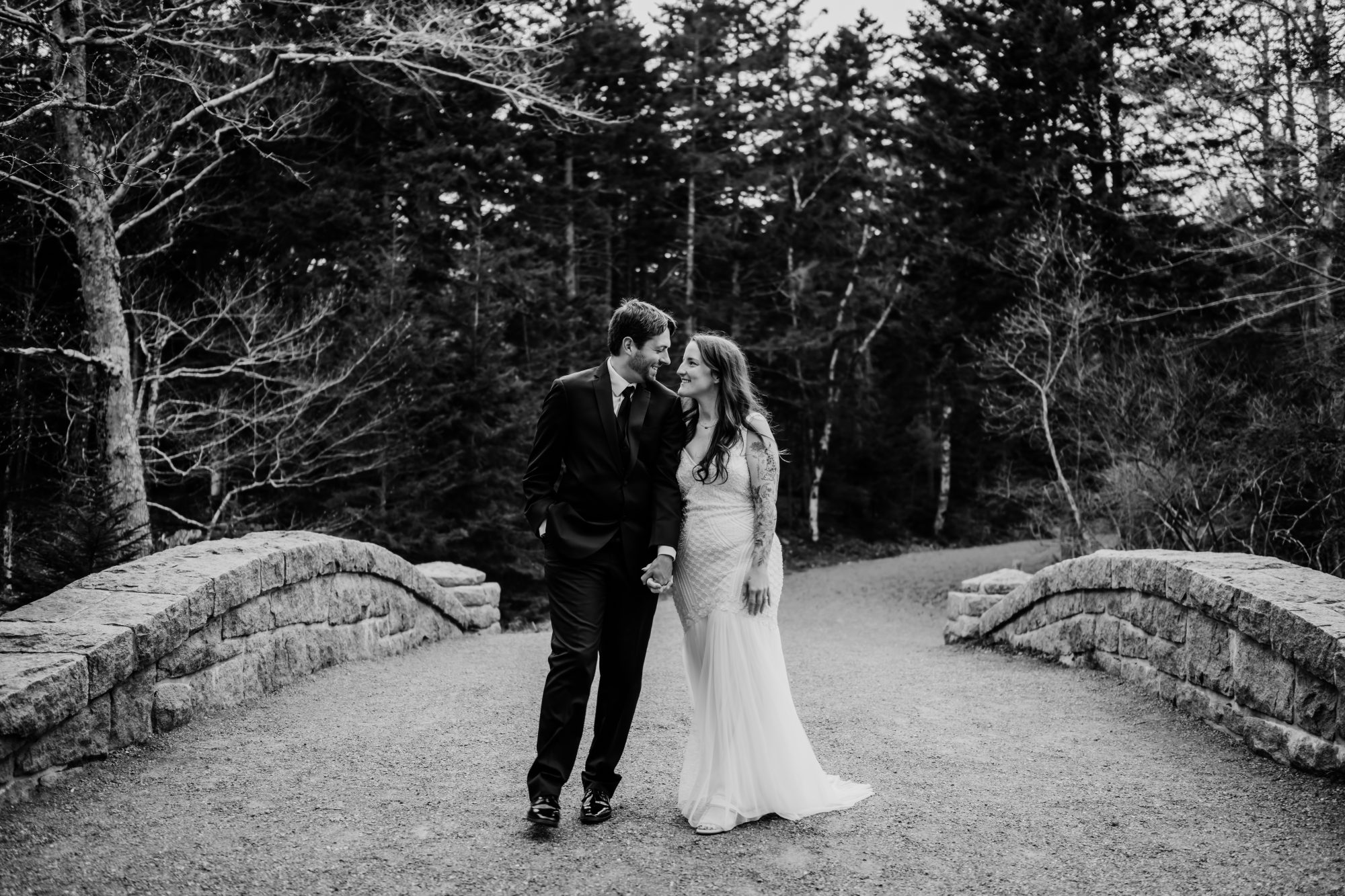 Black and white portrait of an eloping couple walking hand in hand across a stone bridge in Acadia National Park surrounded by evergreen forest