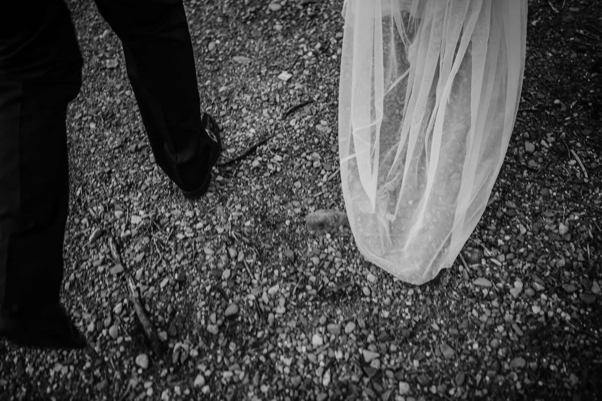 Artistic black and white detail photo of a bride’s flowing wedding dress and groom’s shoes walking along a gravel path during an Acadia elopement