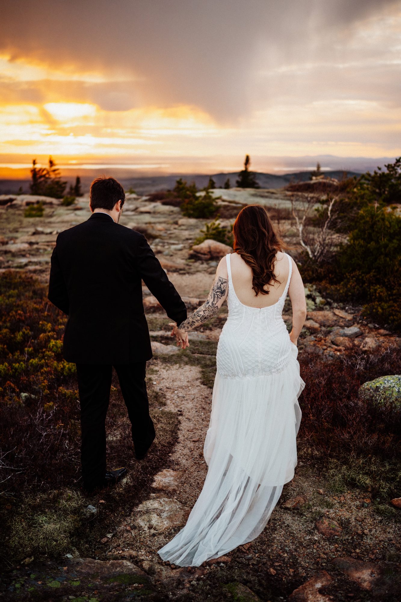 Bride and groom walking hand in hand toward a glowing sunset during their mountaintop elopement in Acadia National Park