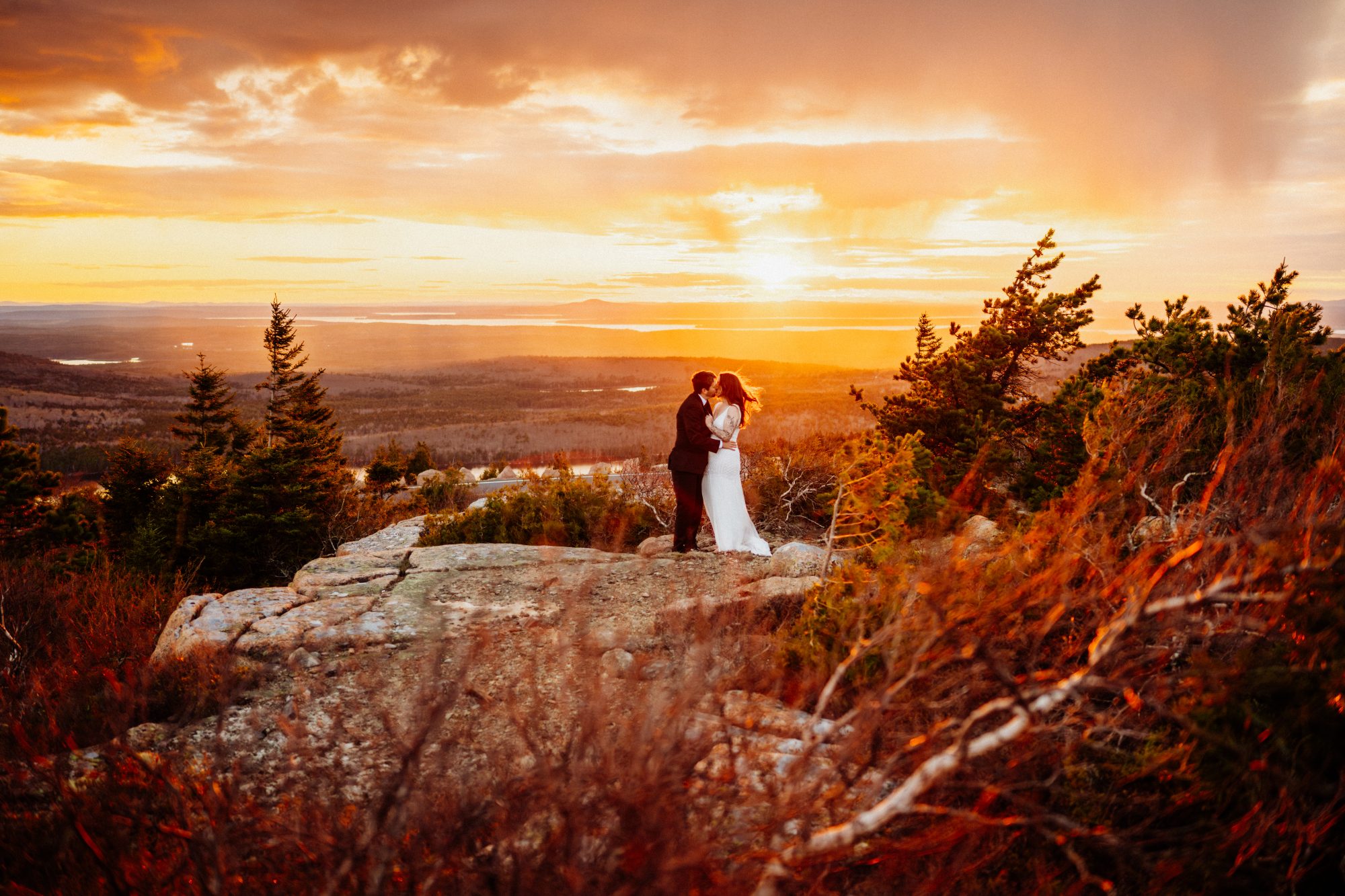 Wide scenic elopement photograph of a couple kissing on a cliffside at sunset in Acadia National Park with dramatic mountain views and glowing sky