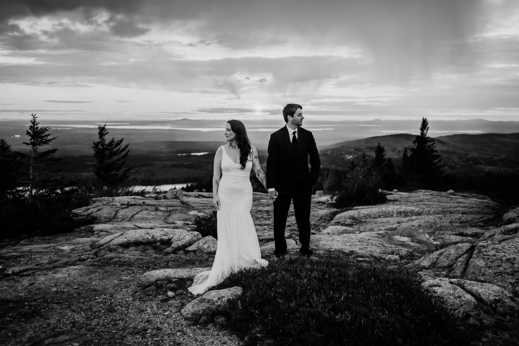 Black and white elopement portrait of a couple holding hands on a mountain summit in Acadia National Park during a dramatic sunset