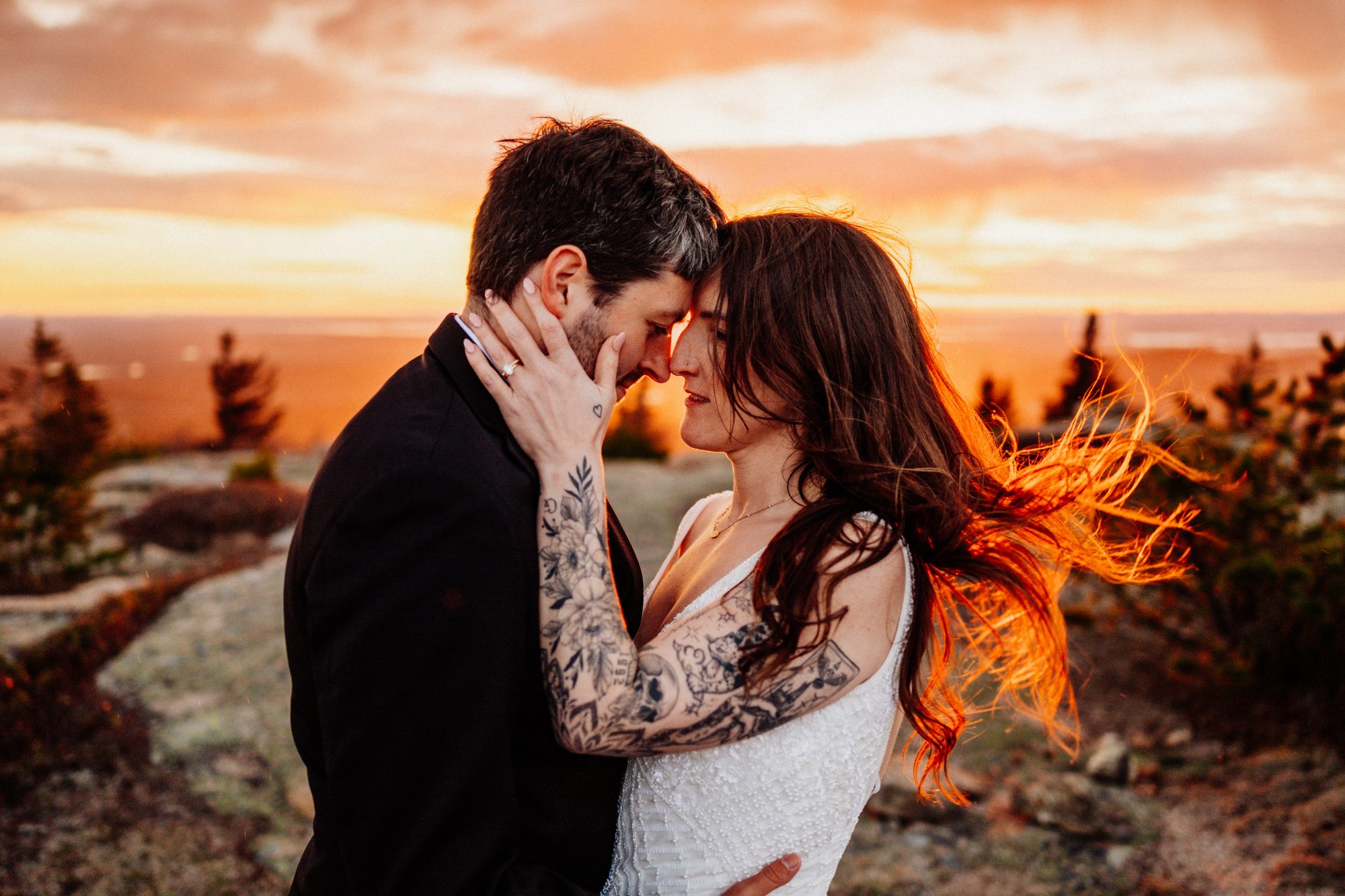 Moody and romantic Acadia National Park elopement photo with glowing sunset light and tattooed bride embracing groom on a mountain overlook